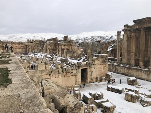 wide-angle view of the ruins of Baalbek with modern apartment buildings and snow-covered mountains in the background. the ruins consist of a standing temple with columns to the right and a one-story platform at center and left, with large blocks strewn about. there is a thin layer of snow on the blocks and tourists walking around.