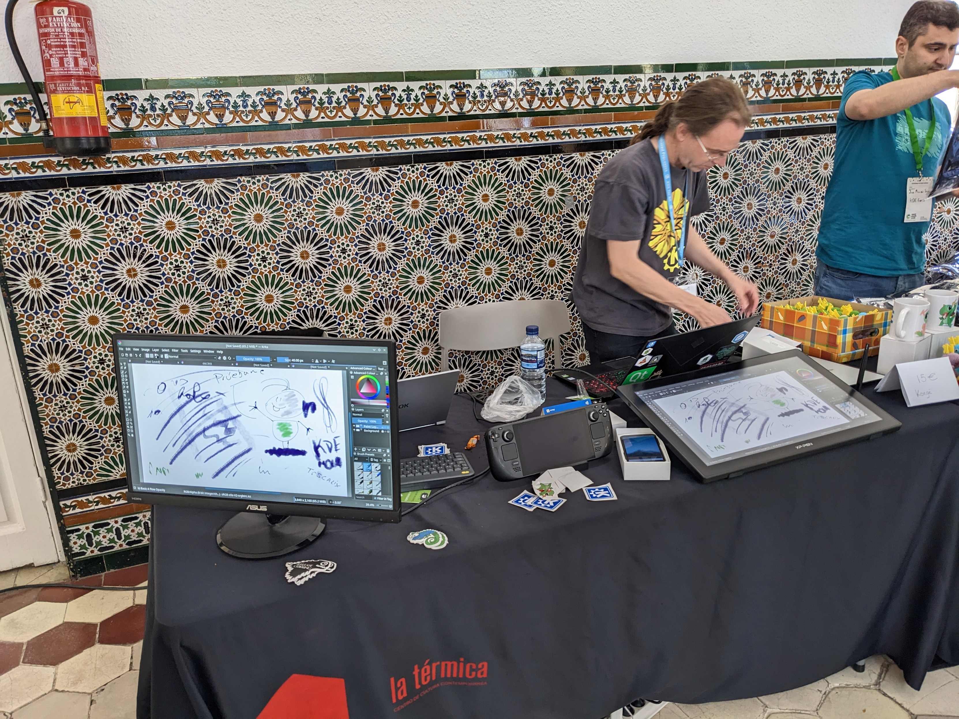 This photo shows the KDE booth at OpenSouthCode 2023. On the table there from left to right a monitor showing pictures drawn with Krita, A Steam Deck, a Pinephone, and a drawing tablet. An assortment of stickers lay scattered around the devices.