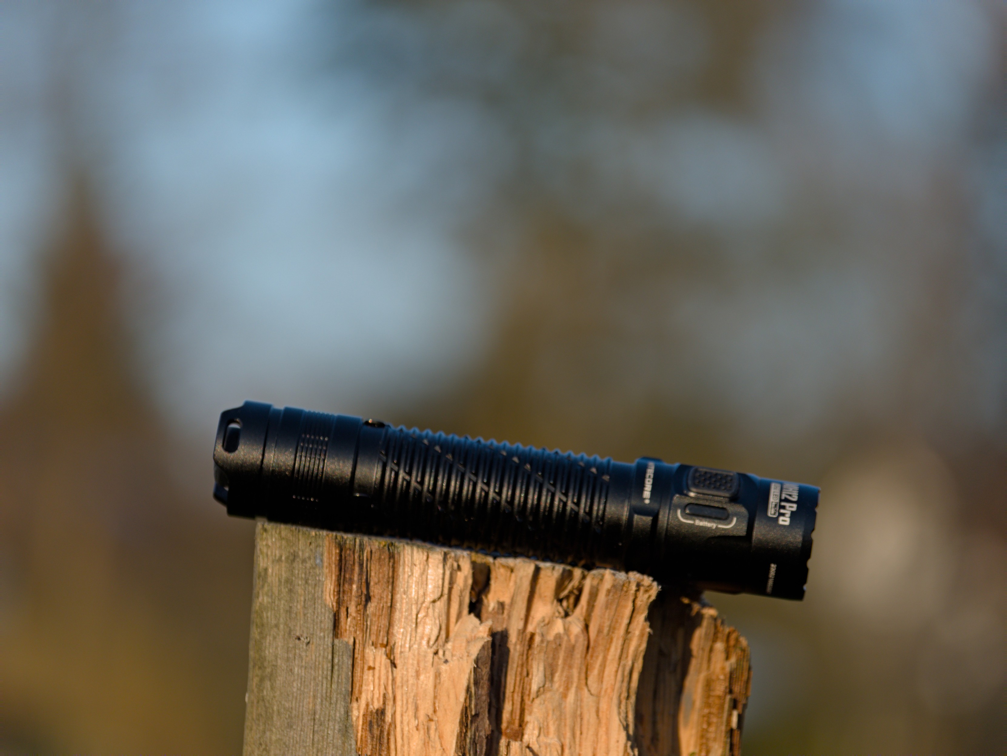 A Nitecore MH12 Pro flashlight on top of a wooden post, with a blurred background