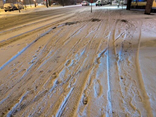 Ein Geh- und Radweg vor einigen Geschäften. Es ist noch vor Sonnenaufgang. Im frischen Schnee sieht man einzelne Fußabdrücke und Spuren von Fahrradreifen. Der Schnee ist großflächig von Autoreifen platt gedrückt.
